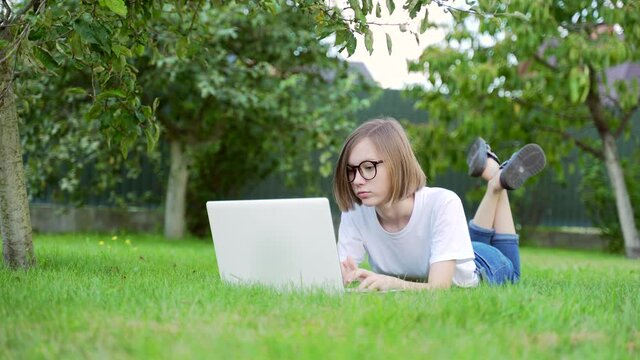 young woman lying on the lawn with a laptop. Girl student in the park on the grass works and studies. Female blonde in a white T-shirt and jeans on a sunny day. Distance learning. Relax in nature