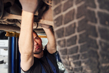 Man in work uniform standing under car and repairs it indoors. Conception of automobile service