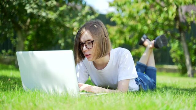 young woman lying on the lawn with a laptop. Girl student in the park on the grass works and studies. Female blonde in a white T-shirt and jeans on a sunny day. Distance learning. Relax in nature