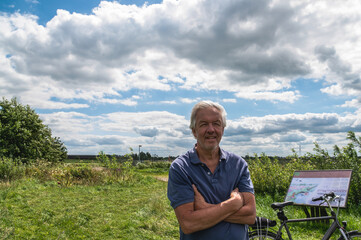 Casual dressed active elderly man stands next to his bicycle at an information board in a typical Dutch landscape with green meadows and Ruisdael clouds.