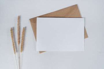 Blank white paper on brown paper envelope with bristly foxtail dry flower. Mock-up of horizontal blank greeting card. Top view of Craft paper envelope on white background. Flat lay minimalism style.
