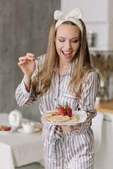 Portrait of a beautiful young woman on the background of the kitchen.A girl in striped pajamas holds a fork and a plate of waffles for Breakfast