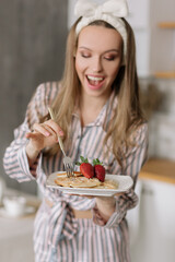 Portrait of a beautiful young woman on the background of the kitchen, having Breakfast in the kitchen.A girl in striped pajamas holds a fork and a plate of waffles for Breakfast