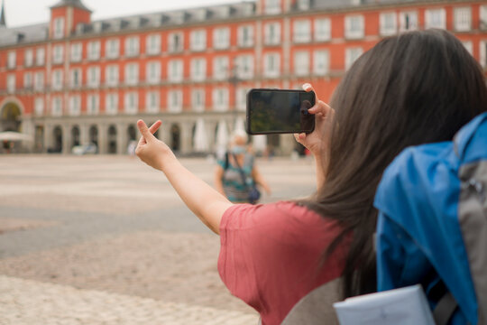 new normal holidays travel in Europe - young beautiful and happy Asian Korean tourist woman with face mask and backpack taking pictures with mobile phone