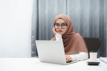 Young muslim business woman wearing brown hijab working with laptop computer in her house. Look at camera.