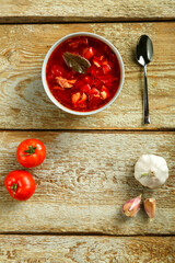 A plate of borscht with chicken with a spoon on a wooden table next to garlic and tomatoes.