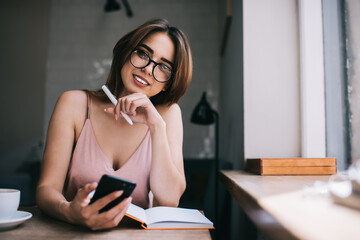 Happy young woman surfing mobile phone and writing info in organizer in modern room