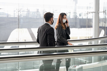 Business woman wearing face mask using smartphone while standing on escalator with businessman nearby, business trip and stay safe concept.