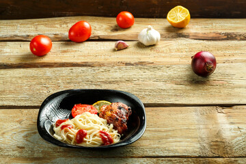 Black plate of spaghetti with baked chicken on a wooden table near vegetables.