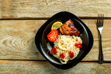 Black plate of spaghetti with baked chicken with a fork on a wooden table.