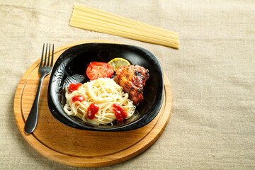 Black plate of spaghetti with baked chicken on a stand with a fork next to uncooked pasta.