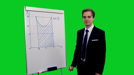 Young male student in suit writing mathematical formula on board against green screen background,...