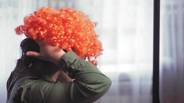 A woman in front of a mirror puts on a clown wig with red curls.