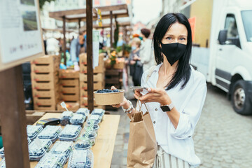 Brunette slim attractive woman choosing and buying blueberries in her container durring summer farmer market outdoor in city street.