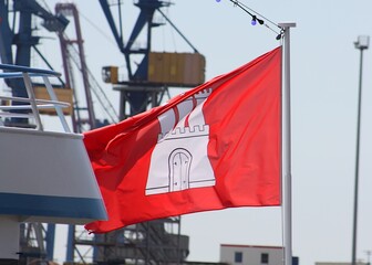 Blick auf die Flagge der freien und Hansestadt Hamburg mit Hafenkränen im Hintergrund