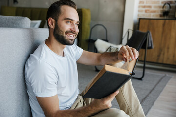 Image of young happy man smiling and reading book while sitting on couch