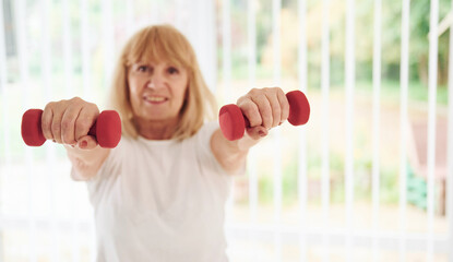 Positive senior woman in sportive clothes indoors at daytime doing exercises with dumbbells