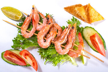 Shrimps on skewers on lettuce leaves with cucumber and tomato slices on a white surface.