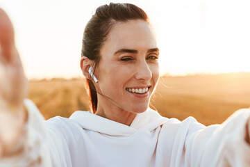 Image of happy sportswoman in earphones winking and taking selfie photo