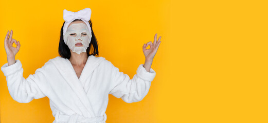 young woman in a white bathrobe on a yellow background. Cloth cosmetic mask on the face. skin rejuvenation and cleansing concept. meditation and calmness.