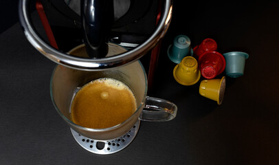 A delicious coffee with foam made by a capsule coffee maker and several colored capsules next to it.