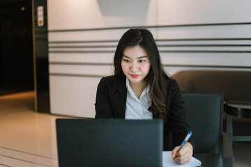 Beautiful young asian woman in suit using laptop and writing note