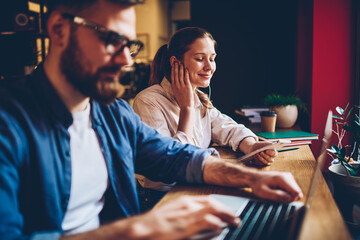 Young female teenager enjoying favourite music playlist listening via application on cellular gadget and electronic earphones, hipster girl spending time for audio book in cafeteria with male friend