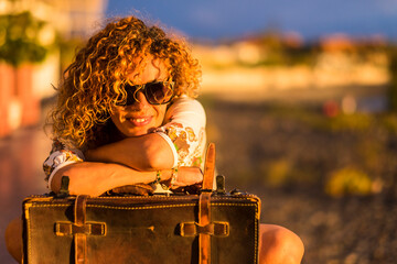 Beautiful curly hair young woman enjoy and relax outdoor with old luggage and smile - concept of travel and happy lifestyle with cheerful people - sunset time and orange colors