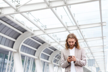 Portrait of handsome curly businessman in casual wear holding smartphone and smiling.Successful manager using mobile phone apps,texting message,browsing internet,looking at phone near business center.