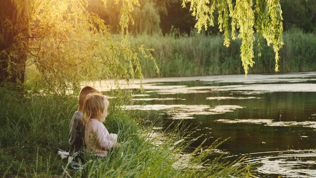 Side view of a couple of children, a cute little boy and a girl, are sitting on the grass by a small lake at sunset, talking by throwing pieces of bread into the water at sunset