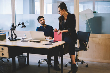 Obraz premium Attractive experienced brunette manager in spectacles sitting on wooden table and explaining main concepts of work to new talented male colleague.Coworkers communicating with each other indoors