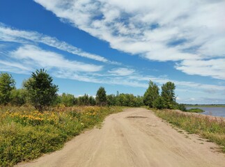 road near the bank of the river among the greenery of grass and trees on a sunny day against the backdrop of a blue sky with clouds