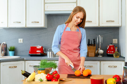 Blonde Young Woman In Apron Cutting Oranges At Her Kitchen