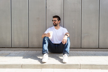 Bearded man with sunglasses sitting on ground while holding a take away coffee