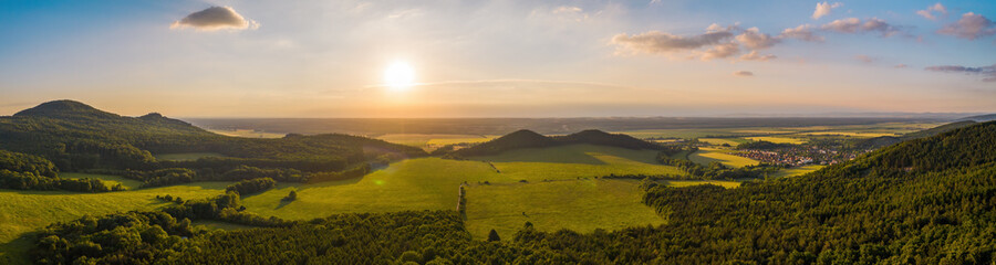 Summer nature scenery at sunset with green forests and meadow and blue sky. Panoramic aerial view on hills with orange sun shining above horizon with copy space.