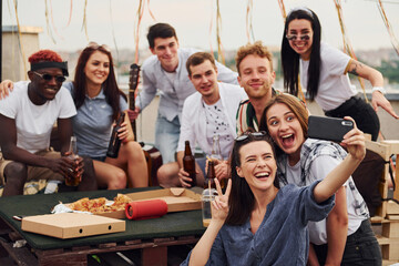 Girl making selfie. With delicious pizza. Group of young people in casual clothes have a party at rooftop together at daytime