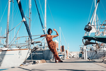 Happy and cheerful young black woman say ahllo standing on the dock with lot of boats around - concept of travel and summer holiday vacation for people and tourism