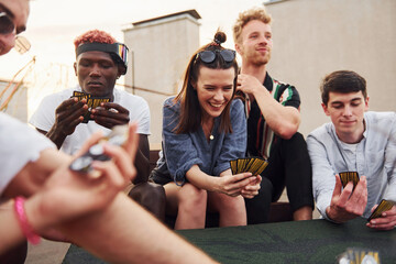 Sitting by table and playing card game. Group of young people in casual clothes have a party at rooftop together at daytime