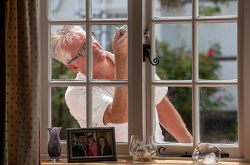 Hampshire, England, UK. 2020. Painter decorator painting small windows on a rural house viewed from inside the home.