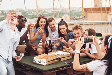Making photo. Delicious pizza. Group of young people in casual clothes have a party at rooftop together at daytime