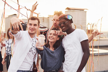 Standing together and taking selfie. Group of young people in casual clothes have a party at rooftop together at daytime