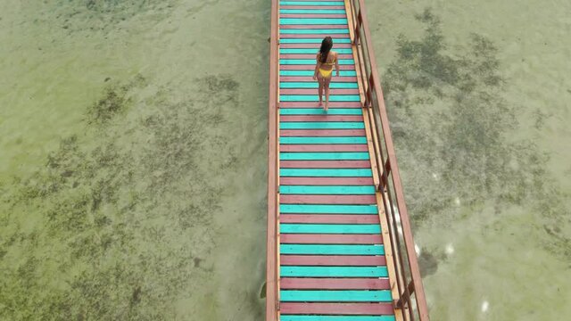 aerial back view drone follows beautiful sexy brunette woman in yellow bikini walking on wooden bridge or pier in clear sea water