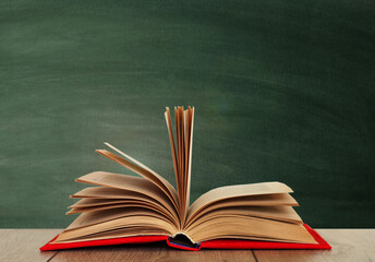Open book textbook in a red cover on a wooden desk on the background of a green school chalkboard. Back to school distance home education.Quarantine concept of stay home.