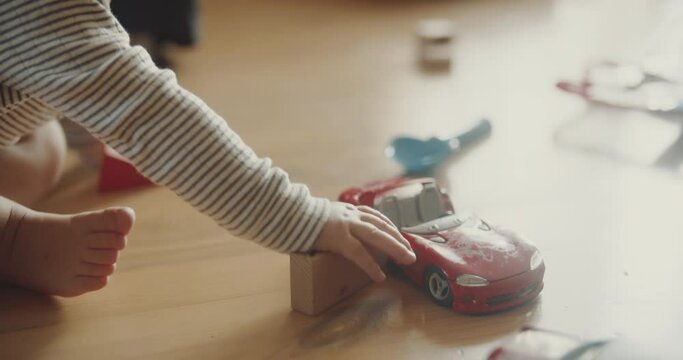 View of childs hands playing red car models and wooden blocks on the floor. Room filled with morning sunlight. Tiny fingers with toys.