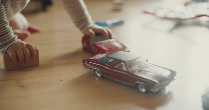 View of childs hands playing red car models and wooden blocks on the floor. Room filled with morning sunlight. Tiny fingers with toys.