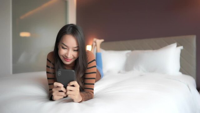 Young asian woman laying on bed and texting on smartphone with smile on her face , full frame slow motion