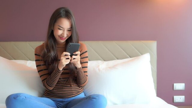 Happy beautiful asian female texting and chatting on mobile phone in her bedroom while sitting, full frame slow motion