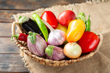Various vegetables in a basket on a brown wooden table. Lots of raw vegetables in the basket. Eggplant, tomatoes, garlic, sweet peppers, onions on a wooden table. Healthy food concept