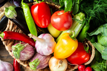 Various vegetables in a basket on a brown wooden table. Lots of raw vegetables in the basket. Eggplant, tomatoes, garlic, sweet peppers, onions on the table. Top view with space for text. Healthy food