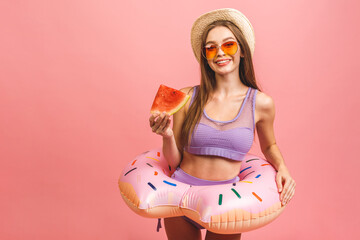 Portrait of a happy young woman dressed in swimsuit jumping and holding swim inflatable ring isolated over pink background. Holding watermelon.